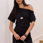 A woman models a black off-the-shoulder short-sleeve mini dress, styled with a black leather hobo bag, black sunglasses, a black and white polka dot bandana, and a silver pendant necklace, standing in an indoor setting with white walls and wooden stairs.