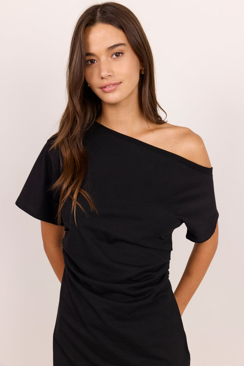 A woman modeling a black off-the-shoulder short-sleeve dress against a plain light beige studio background.