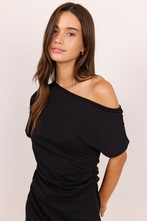 A woman modeling a black off-the-shoulder short-sleeve top against a plain light beige studio background.