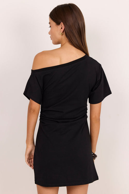 A woman wearing a black off-the-shoulder short-sleeve mini dress, shown from the back against a plain light beige studio background.