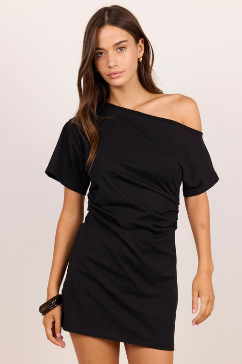 A woman modeling a black off-the-shoulder short-sleeve mini dress against a plain light beige studio background.