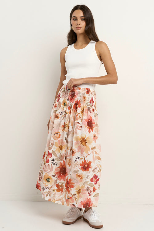 Woman wears a floral maxi skirt with a tiered silhouette, elastic waist, and white ribbed tank top with sneakers.