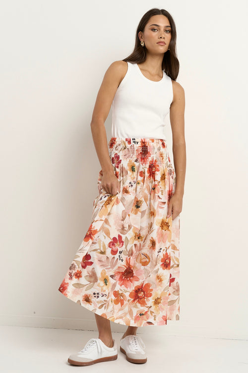 Woman wears a floral maxi skirt with a tiered silhouette, elastic waist, and white ribbed tank top with sneakers.