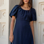 Woman wearing a navy puff-sleeve maxi dress with a gathered scoop neckline and bias-cut silhouette, styled with white sandals in a light architectural setting.







