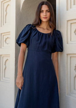 Woman wearing a navy puff-sleeve maxi dress with a gathered scoop neckline and bias-cut silhouette, styled with white sandals in a light architectural setting.







