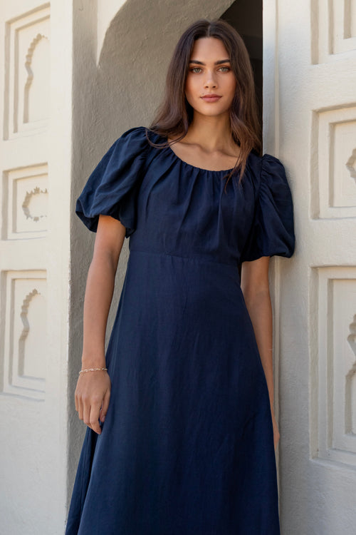 Woman wearing a navy puff-sleeve maxi dress with a gathered scoop neckline and bias-cut silhouette, styled with white sandals in a light architectural setting.







