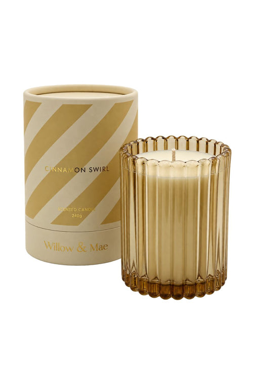 A Willow & Mae candle in a ribbed amber glass holder next to its cylindrical packaging box, which features diagonal stripes in beige and gold with the scent name 'Cinnamon Swirl'.