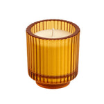 An amber glass candle holder with a ribbed fluted design, containing a lit cream-colored candle, isolated on a white background.