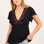 A woman modeling a black short-sleeve top with a lace V-neckline and ruffled sleeves, paired with light-wash blue jeans, against a plain off-white studio background.