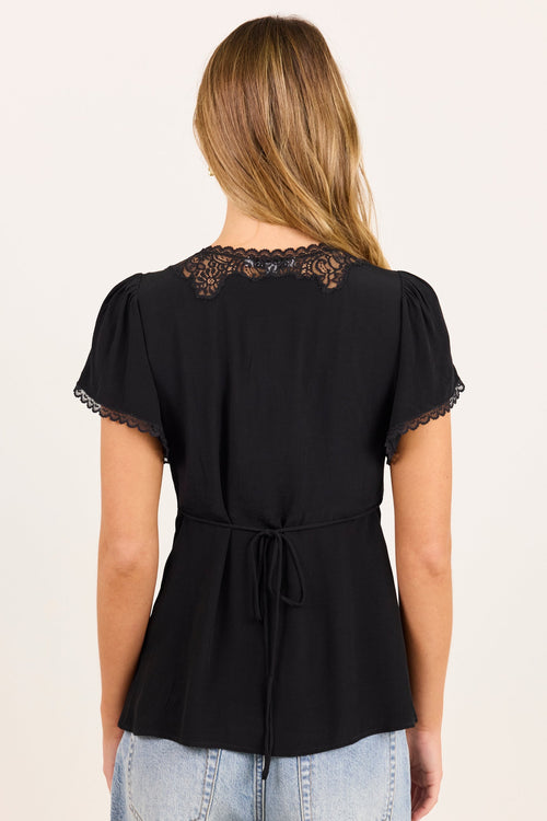 A woman viewed from the back wearing a black short-sleeve peplum top with lace detailing at the neckline and a drawstring tie at the waist, paired with light blue jeans against a plain off-white background.