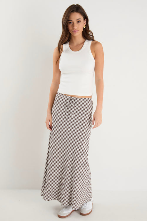 Woman wearing a brown and white gingham print maxi skirt with a tie waist and bias cut, paired with a fitted white tank and white sneakers.