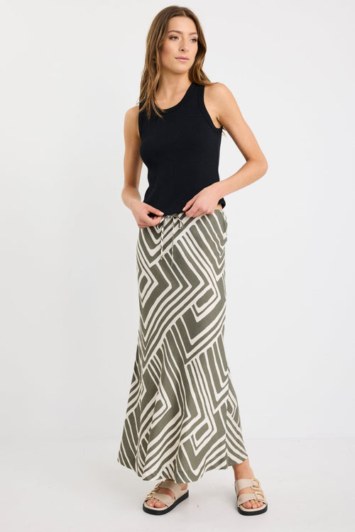 Woman wearing a khaki and ivory geometric print bias-cut maxi skirt with tie waist, paired with a black tank and sandals.