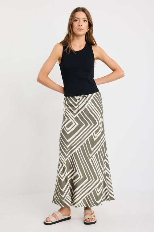 Woman wearing a khaki and ivory geometric print bias-cut maxi skirt with tie waist, paired with a black tank and sandals.