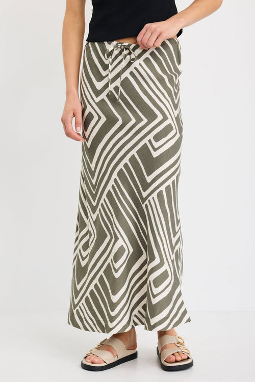 Woman wearing a khaki and ivory geometric print bias-cut maxi skirt with tie waist, paired with a black tank and sandals.