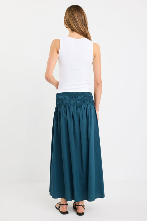 Woman wearing a petrol blue cotton maxi skirt with wide shirred waistband, styled with a white tank and black sandals.