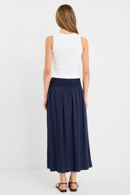 Woman wearing a navy maxi skirt with shirred waistband, styled with a white ribbed tank and black sandals.