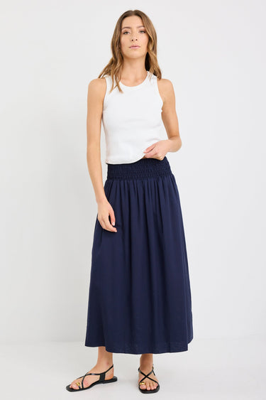 Woman wearing a navy maxi skirt with shirred waistband, styled with a white ribbed tank and black sandals.