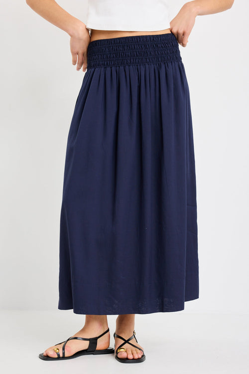 Woman wearing a navy maxi skirt with shirred waistband, styled with a white ribbed tank and black sandals.