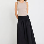 Woman wearing a black cotton maxi skirt with shirred waistband, styled with a taupe ribbed tank and black woven flats.