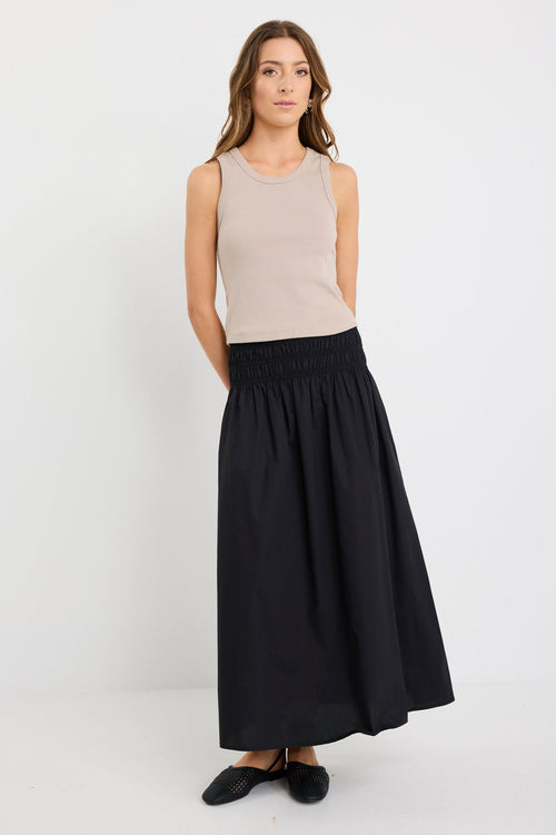 Woman wearing a black cotton maxi skirt with shirred waistband, styled with a taupe ribbed tank and black woven flats.