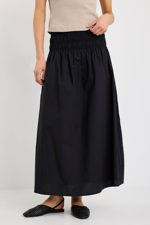 Woman wearing a black cotton maxi skirt with shirred waistband, styled with a taupe ribbed tank and black woven flats.