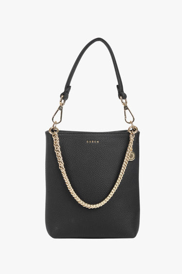 Front-facing view of a black leather bucket bag with a pebbled finish. The structured silhouette is elevated with a bold gold-tone chain draped across the front and matching hardware, including a circular branded charm. A detachable top handle completes the look, with the brand name 'SABEN' embossed in gold near the opening.