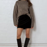 Woman wearing a brown sweater, black skirt, and knee-high boots against a white brick wall.