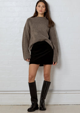 Woman wearing a brown sweater, black skirt, and knee-high boots against a white brick wall.