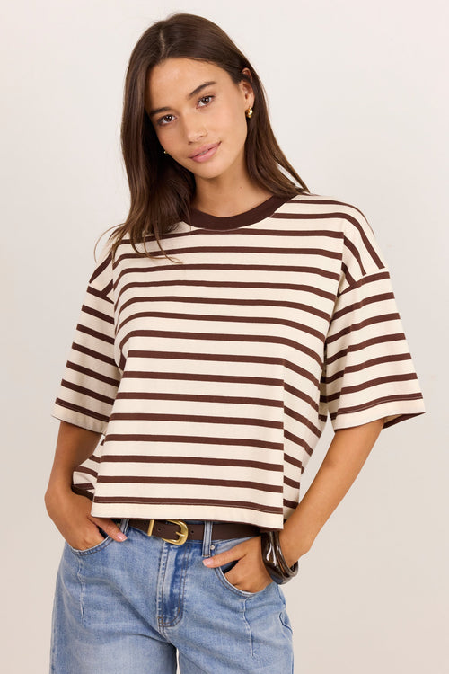 A woman modeling a brown and cream horizontal striped short-sleeve t-shirt with a dark brown crew neck, paired with light-wash blue jeans, against a plain light beige background.