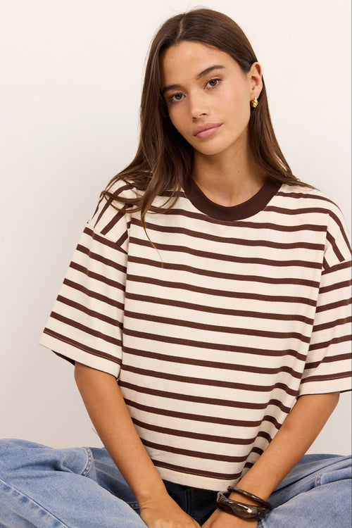 A woman wearing a brown and cream striped short-sleeve t-shirt with a solid brown crew neck, paired with light blue jeans, seated against a plain off-white background.