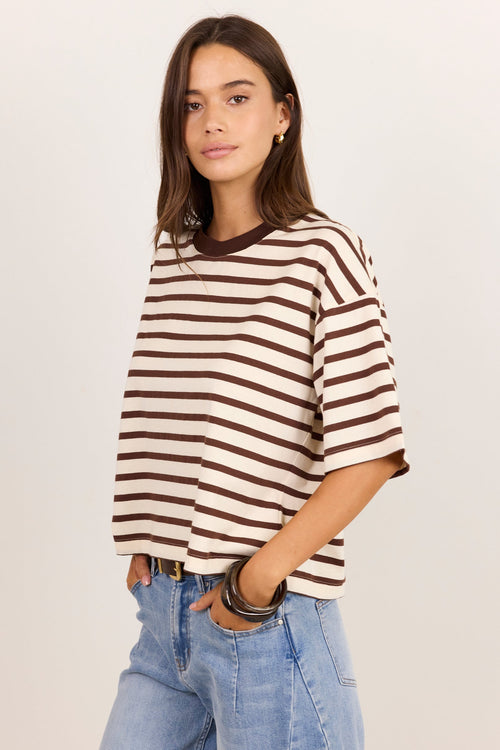 A woman modeling a brown and cream striped short-sleeve knit top with light blue jeans, standing against a plain off-white studio background.