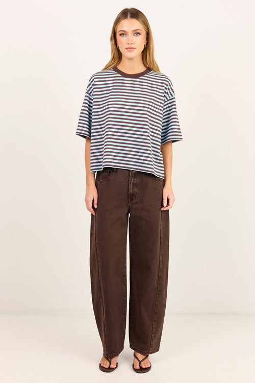 A woman stands against a plain off-white studio background wearing a loose short-sleeve striped t-shirt with navy, blue, and white horizontal stripes, paired with dark brown wide-leg pants and brown sandals.