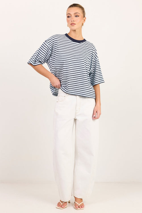 A woman modeling a navy and white horizontally striped short-sleeve t-shirt with white wide-leg jeans and beige sandals, standing against a plain off-white studio background.