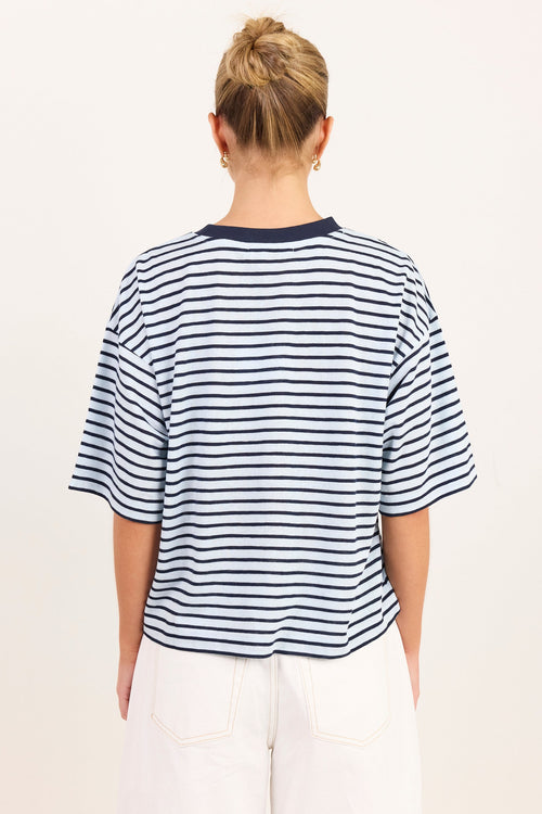A woman viewed from behind wearing a navy and white horizontally striped short-sleeve t-shirt and white pants, set against a plain off-white studio background.