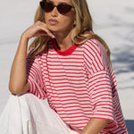 A woman wearing a red and white striped t-shirt and white pants sits on a white tiled poolside deck in a sunny outdoor setting.