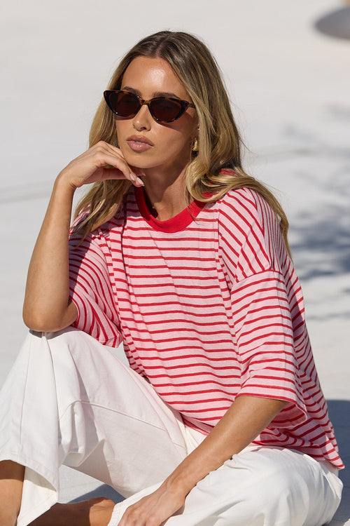 A woman wearing a red and white striped t-shirt and white pants sits on a white tiled poolside deck in a sunny outdoor setting.