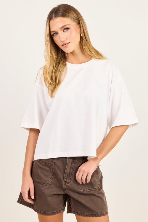 A woman modeling a white short-sleeve crewneck t-shirt and brown denim shorts against a plain off-white studio background.