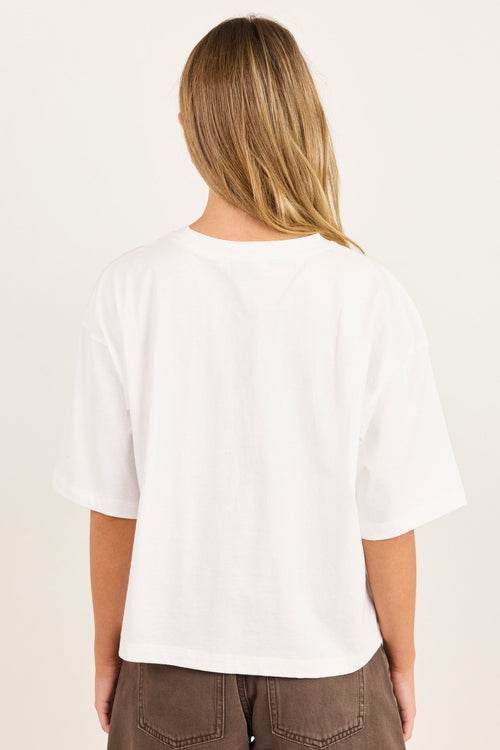 A woman viewed from behind wearing a white short-sleeve crewneck t-shirt and brown pants, set against a plain off-white background.