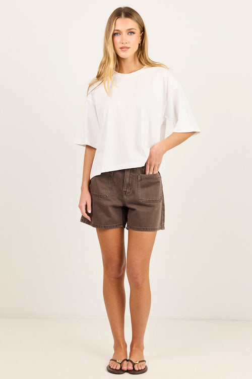 A woman modeling a white short-sleeve t-shirt and brown shorts against a plain off-white studio background.