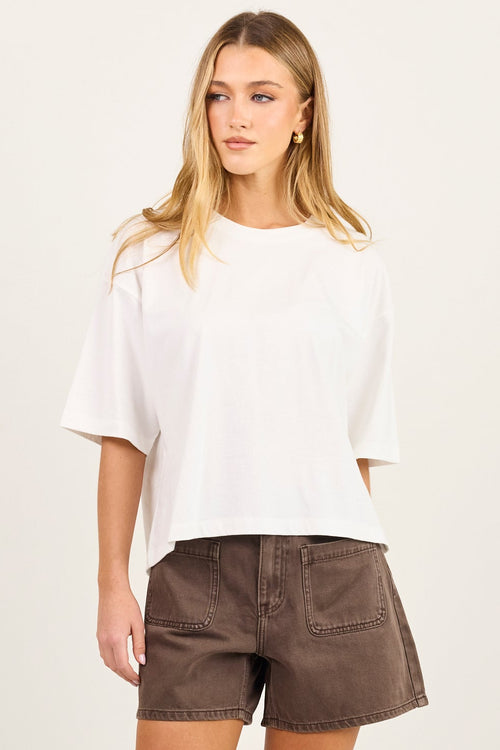 A woman modeling a white short-sleeve crop top and brown shorts against a plain off-white studio background.