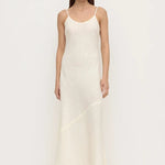 A woman modeling a long, sleeveless white slip dress with thin spaghetti straps and black flat shoes, standing against a plain white studio background.