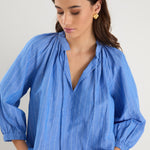 Woman wearing a relaxed-fit, blue pinstripe top with mid sleeves and a split neckline, styled with off-white jeans.