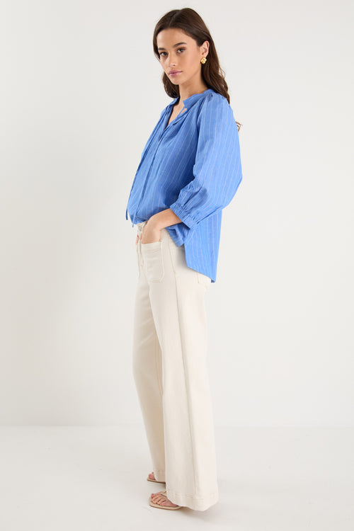 Woman wearing a relaxed-fit, blue pinstripe top with mid sleeves and a split neckline, styled with off-white jeans.