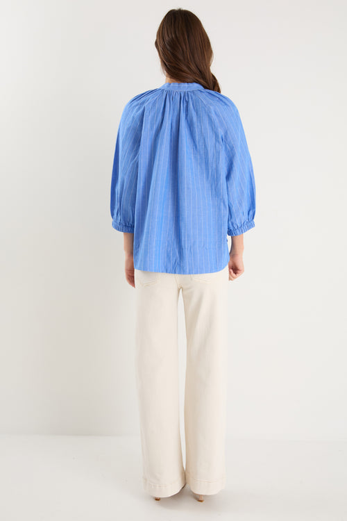 Woman wearing a relaxed-fit, blue pinstripe top with mid sleeves and a split neckline, styled with off-white jeans.