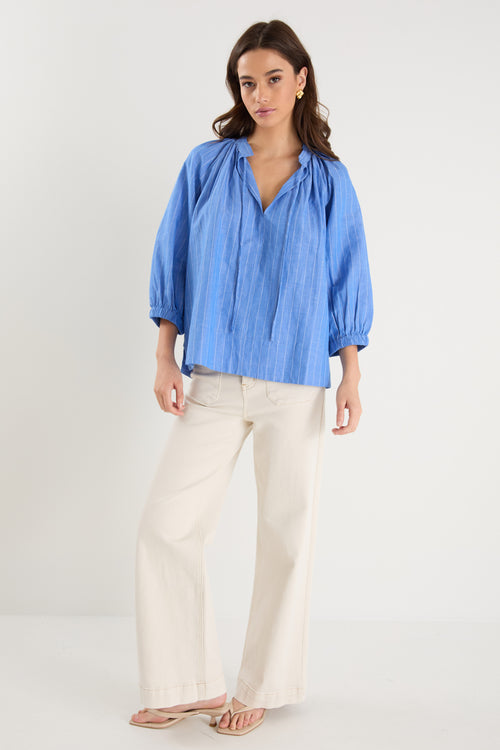 Woman wearing a relaxed-fit, blue pinstripe top with mid sleeves and a split neckline, styled with off-white jeans.