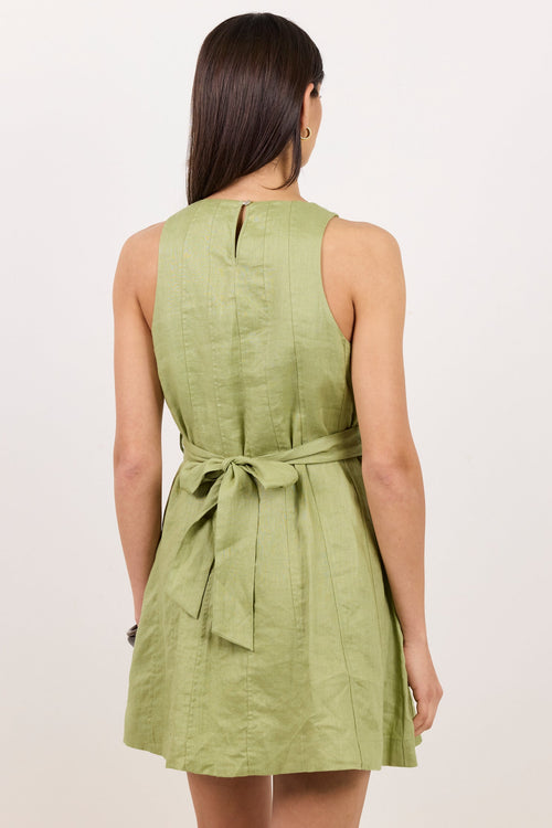 A woman viewed from behind wearing a sleeveless light olive green dress with a tie waist, set against a plain off-white background.
