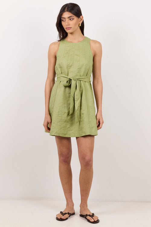 A woman modeling a sleeveless olive green linen mini dress with a tie waist, paired with black flip-flop sandals, against a plain white studio background.