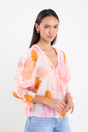 Woman wearing a pink and orange abstract printed short sleeve top with a V-neckline, tie front, and relaxed fit, paired with light blue jeans.