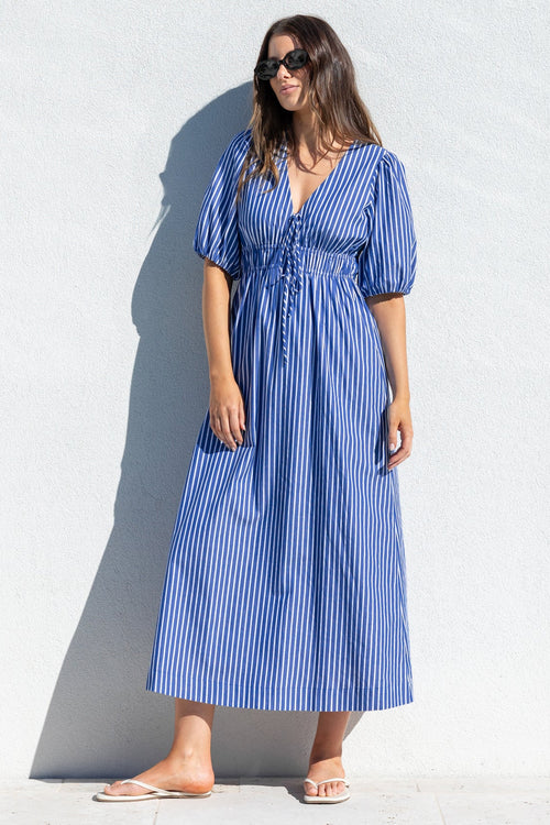 A woman wearing a blue and white vertically striped midi dress with puff sleeves and a V-neckline, standing against a white wall on a sunny day.