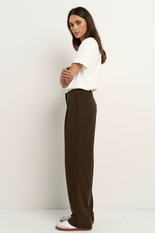 Woman wearing chocolate brown wide leg trousers with a white tee and white sneakers.







